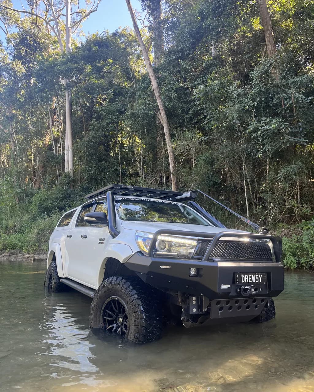 A white Hilux with a bull bar, snorkel and roof rack standing in a clear creek, rainforest behind.
